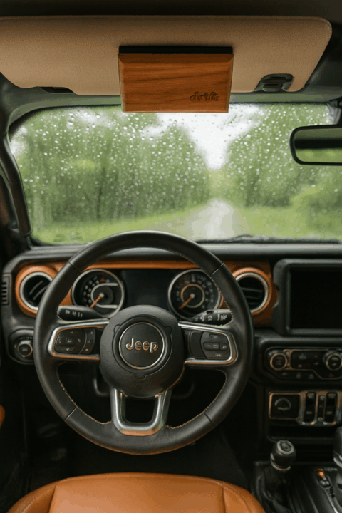 First-person view from inside a Jeep Wrangler with brown leather seats, showing a Drift wood car freshener clipped to the sun visor on a rainy day.