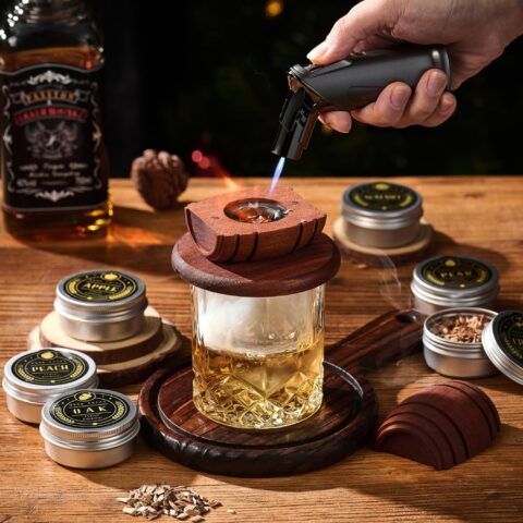 Level Up Your Old Fashioned with a Cocktail Smoker Kit with Torch Hand using a cocktail smoker kit with torch to infuse whiskey in a glass, surrounded by tins of wood chips labeled apple, peach, and oak.