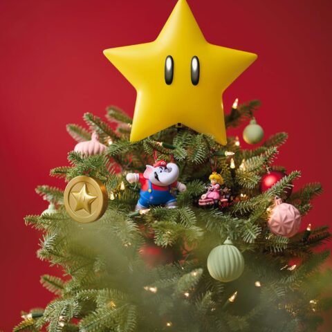 Add Some Nintendo Magic with the Super Mario Star Tree Topper A glowing Super Mario Star tree topper sits on top of a decorated Christmas tree, surrounded by Mario, Princess Peach, and star-themed ornaments against a festive red background.